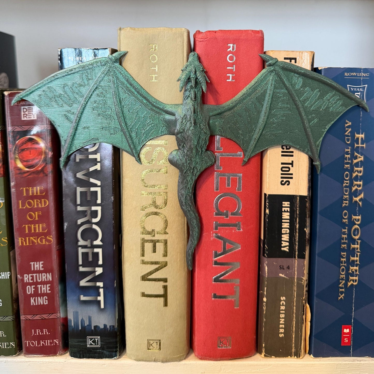 A green dragon book spine hugger with outstretched wings rests between a collection of books. The dragon is detailed with textured wings and a long tail. The books have various colors and titles, creating a fantasy theme.