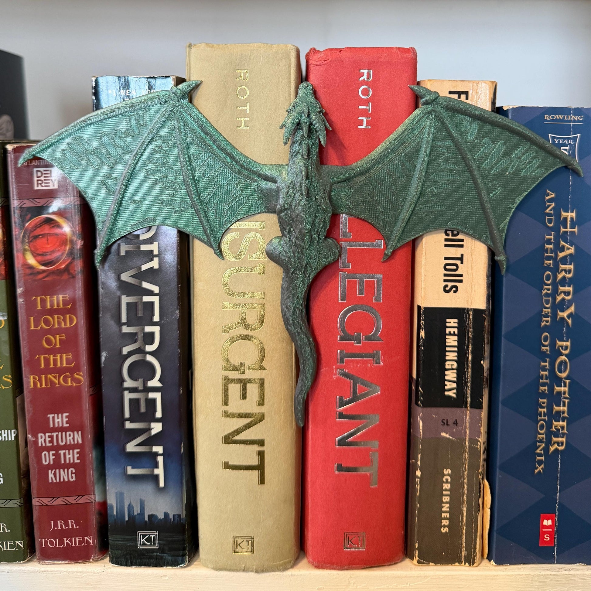 A green dragon book spine hugger with outstretched wings rests between a collection of books. The dragon is detailed with textured wings and a long tail. The books have various colors and titles, creating a fantasy theme.