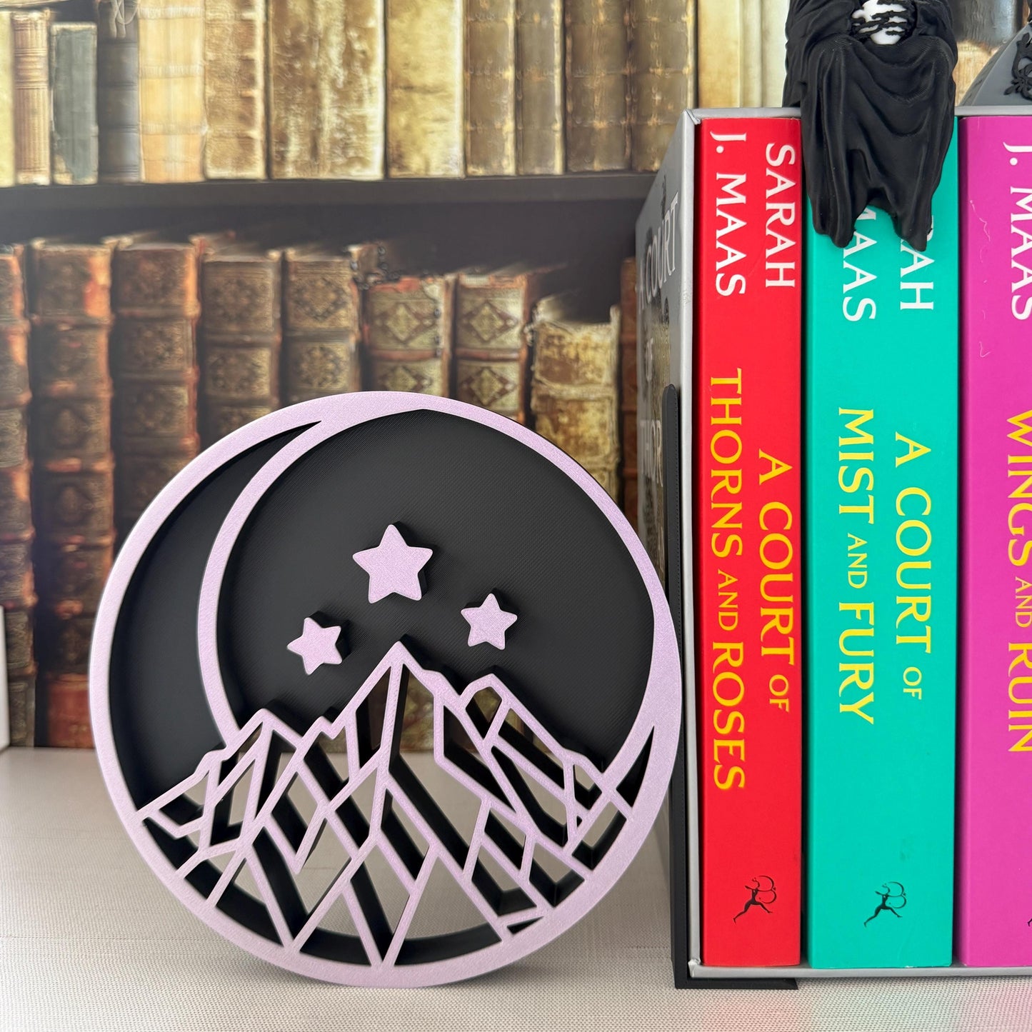 May include: A round, black and lavender bookend featuring a mountain range, crescent moon, and three stars design. The bookend is next to three books with titles by Sarah J. Maas.