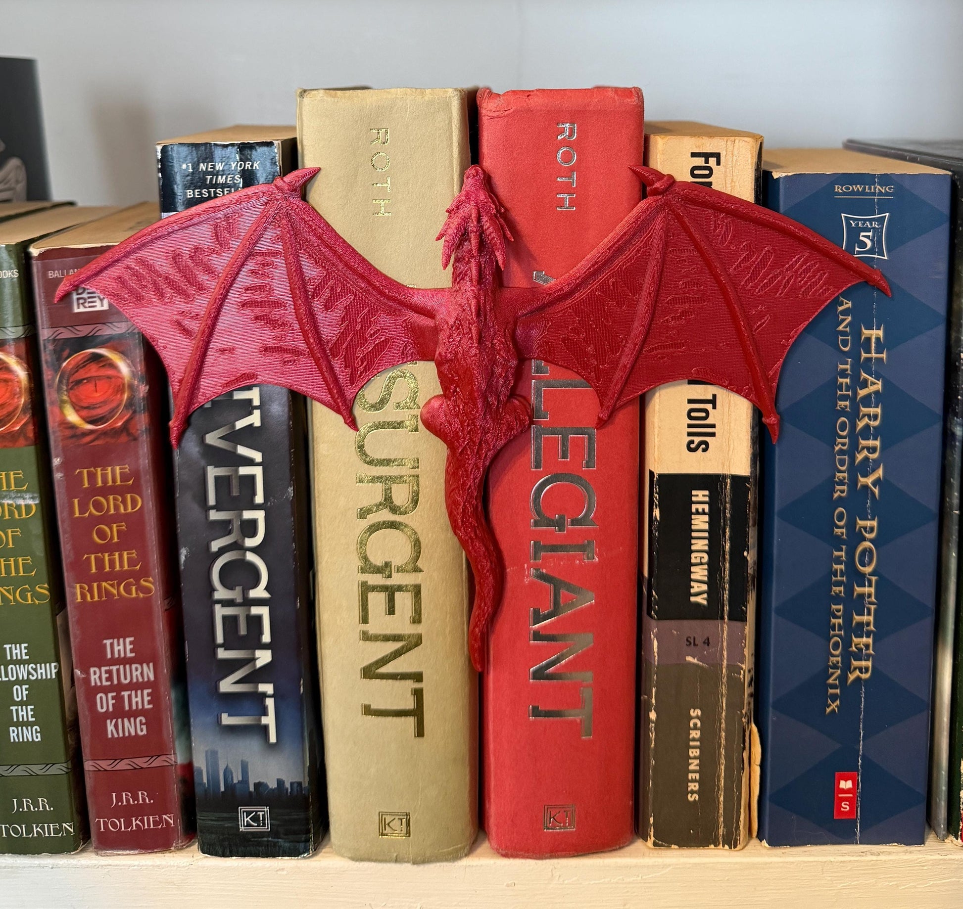 A red dragon book spine hugger with outstretched wings, placed between a row of books. The dragon is positioned to appear as if it is perched on the spines of the books. The books have titles such as "Harry Potter" and "The Lord of the Rings".