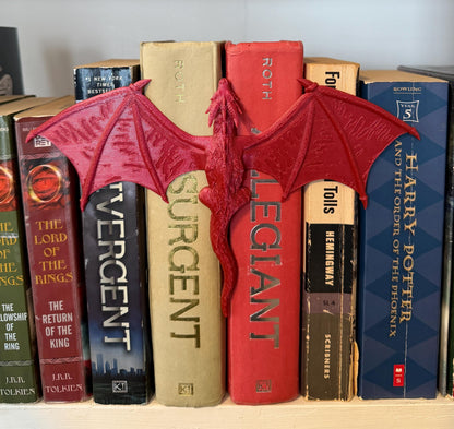 A red dragon book spine hugger with outstretched wings, placed between a row of books. The dragon is positioned to appear as if it is perched on the spines of the books. The books have titles such as "Harry Potter" and "The Lord of the Rings".