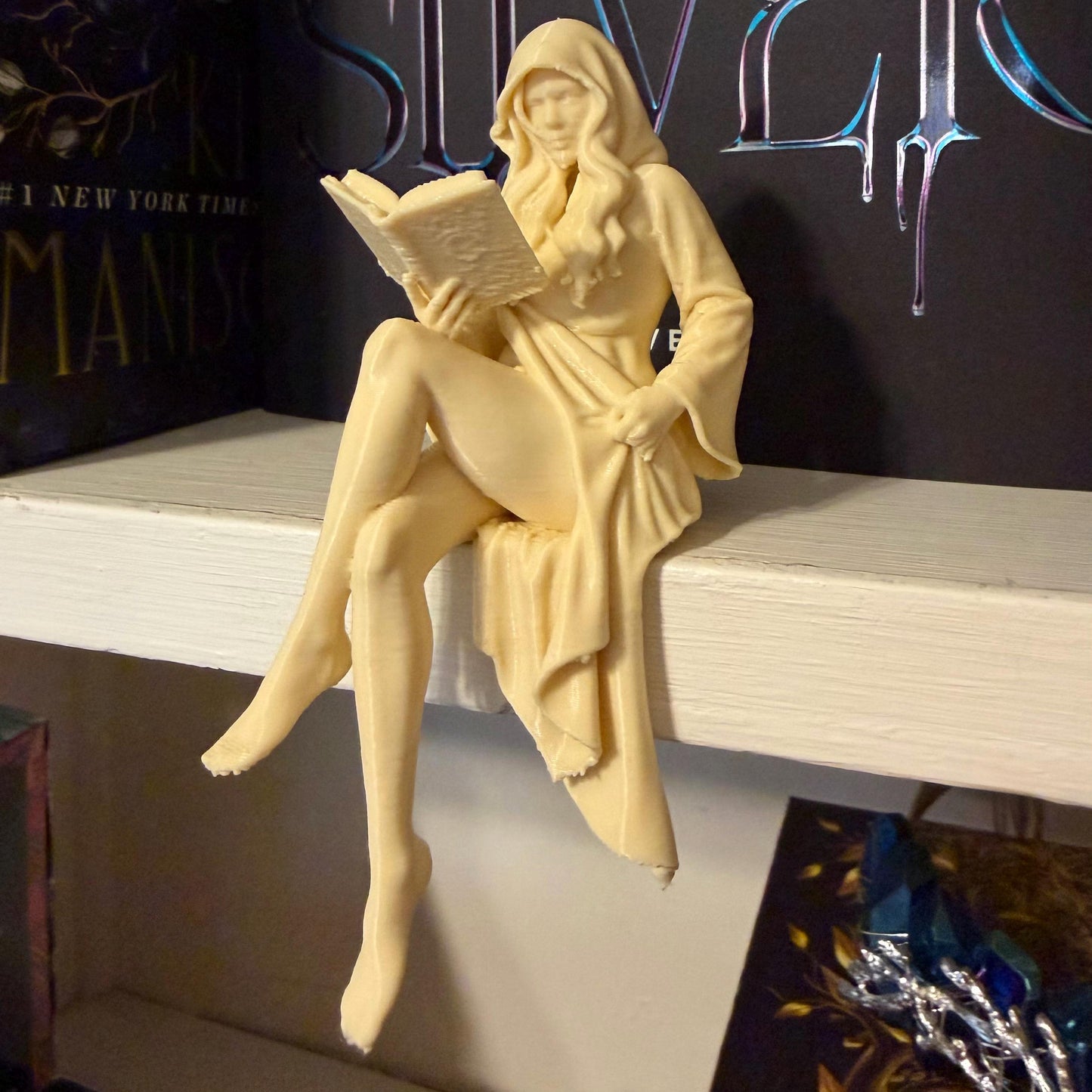 May include: A cream-colored figurine of a woman in a hooded robe, seated and reading a book. The figure is perched on a white shelf, with a book title visible in the background. The figurine has long hair and is holding the book open.