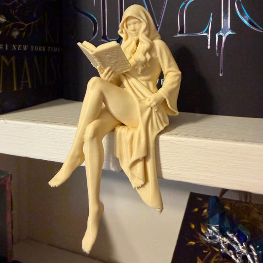 May include: A cream-colored figurine of a woman in a hooded robe, seated and reading a book. The figure is perched on a white shelf, with a book title visible in the background. The figurine has long hair and is holding the book open.