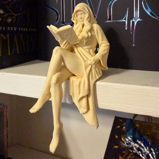 May include: A cream-colored figurine of a woman in a hooded robe, seated and reading a book. The figure is perched on a white shelf, with a book title visible in the background. The figurine has long hair and is holding the book open.