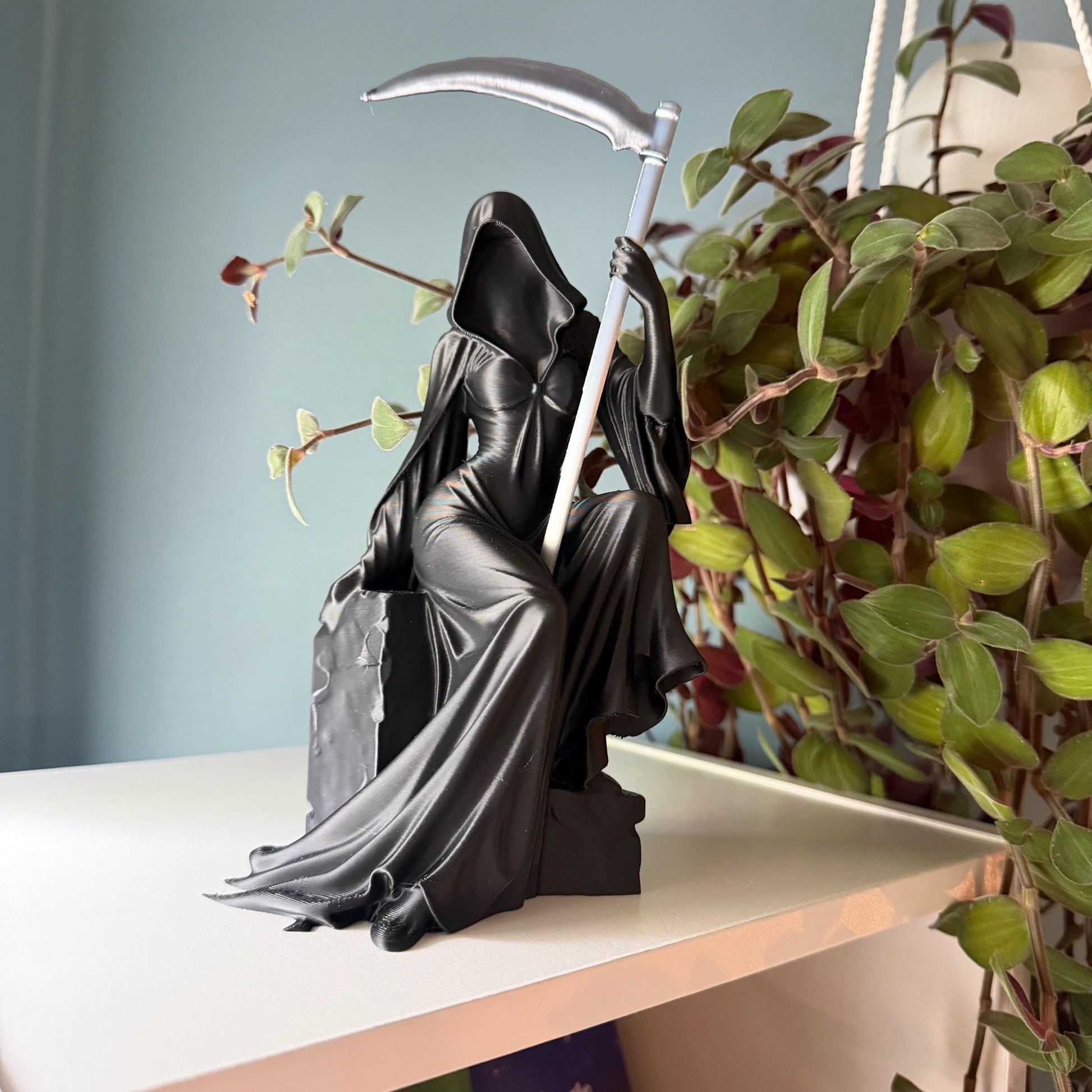 May include: A black Grim Reaper statue with a silver scythe. The figure is seated, wearing a hooded cloak and a flowing gown. The statue is on a white shelf, with green plants in the background.