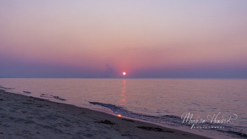 Lake Michigan at sunset, with gentle waves, a smooth sandy shoreline, and a glowing orange sun on the horizon.