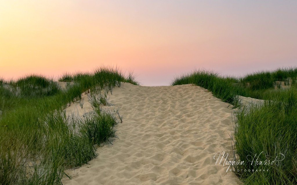Sandy trail flanked by dune grasses under a pastel orange and pink sunset sky.